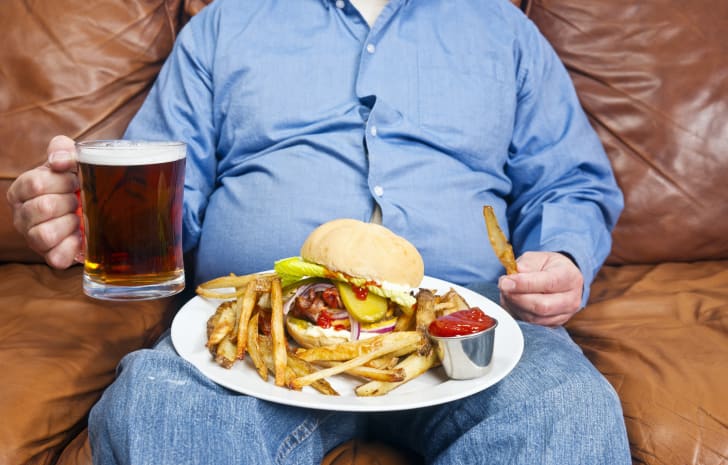 Um homem, mostrado do pescoço para baixo, sentado em um sofá segurando uma cerveja com um hambúrguer e batatas fritas em um prato no colo.